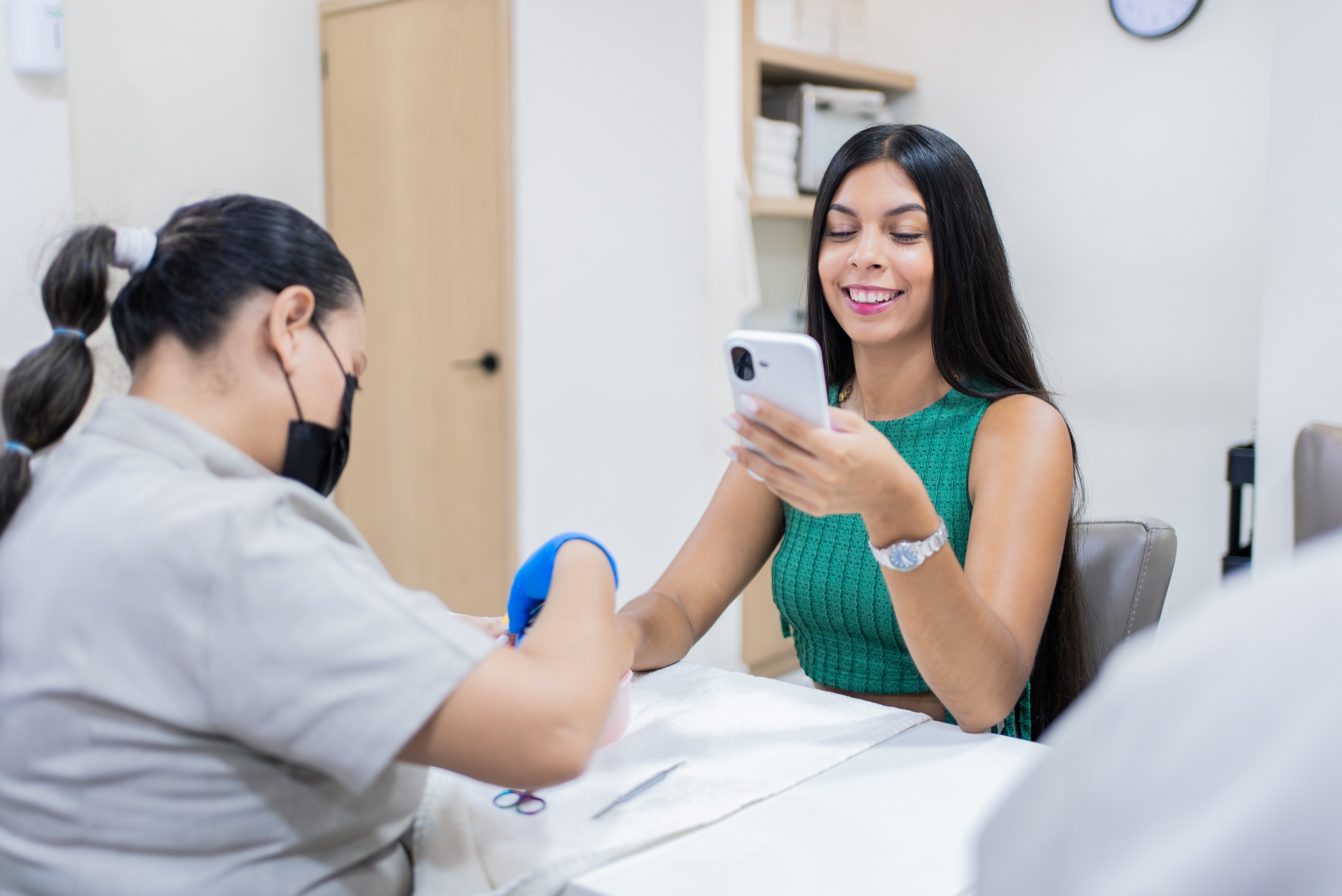 Beautician giving manicure to woman using smartphone in beauty salon